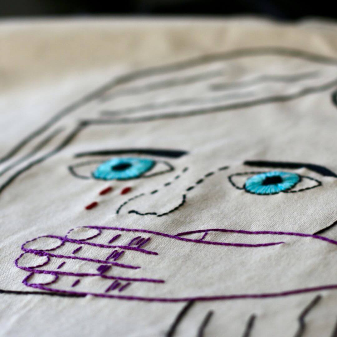 Cloth embroidered with the outline and contours of a women's face in black thread, with blue thread for eyes and another person's hand in purple holding her mouth shut. 