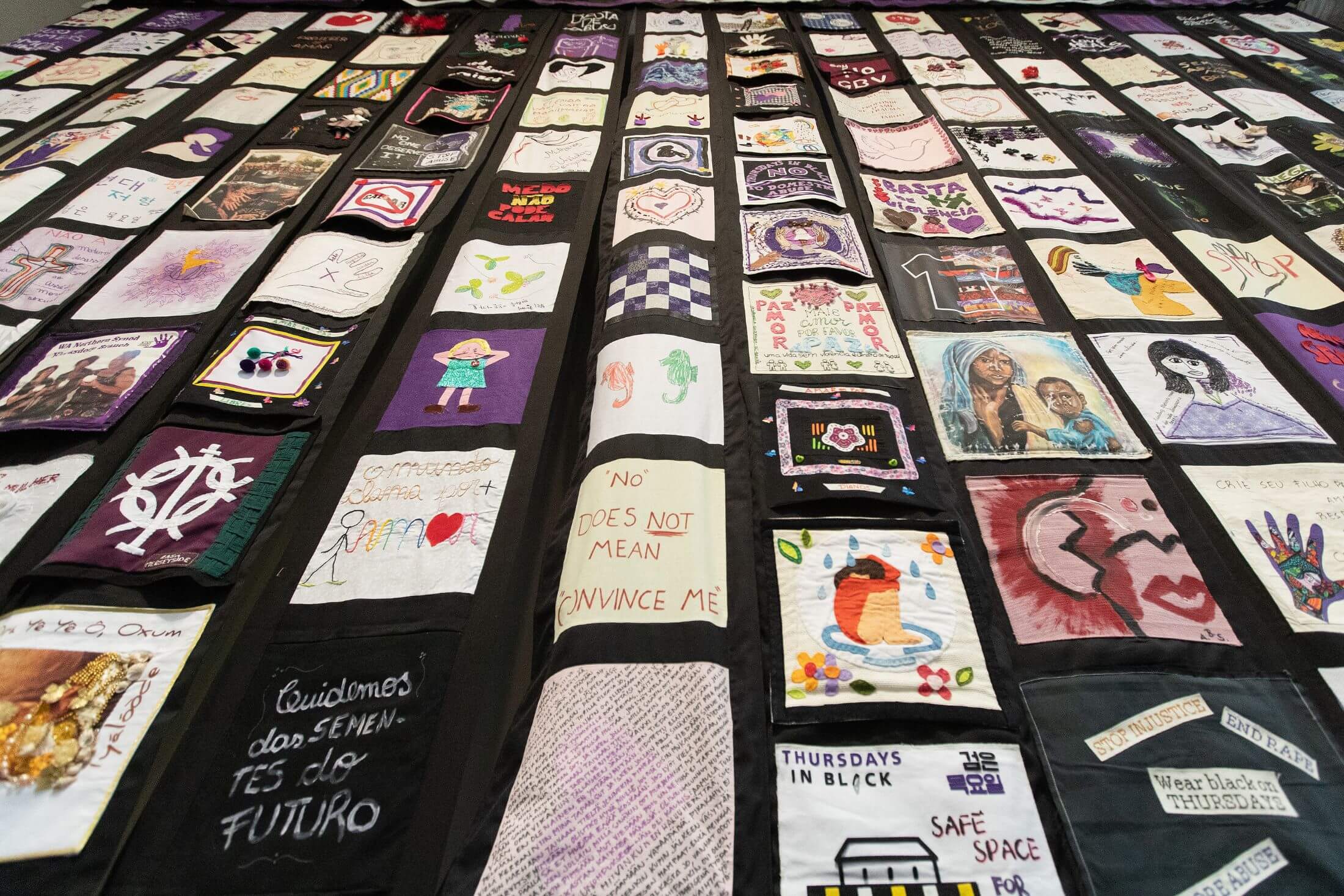 A quilt with fabric blocks depicting girls and women hurt by violence and slogans calling for an end to gender-based violence. Exterior and interior borders are black.