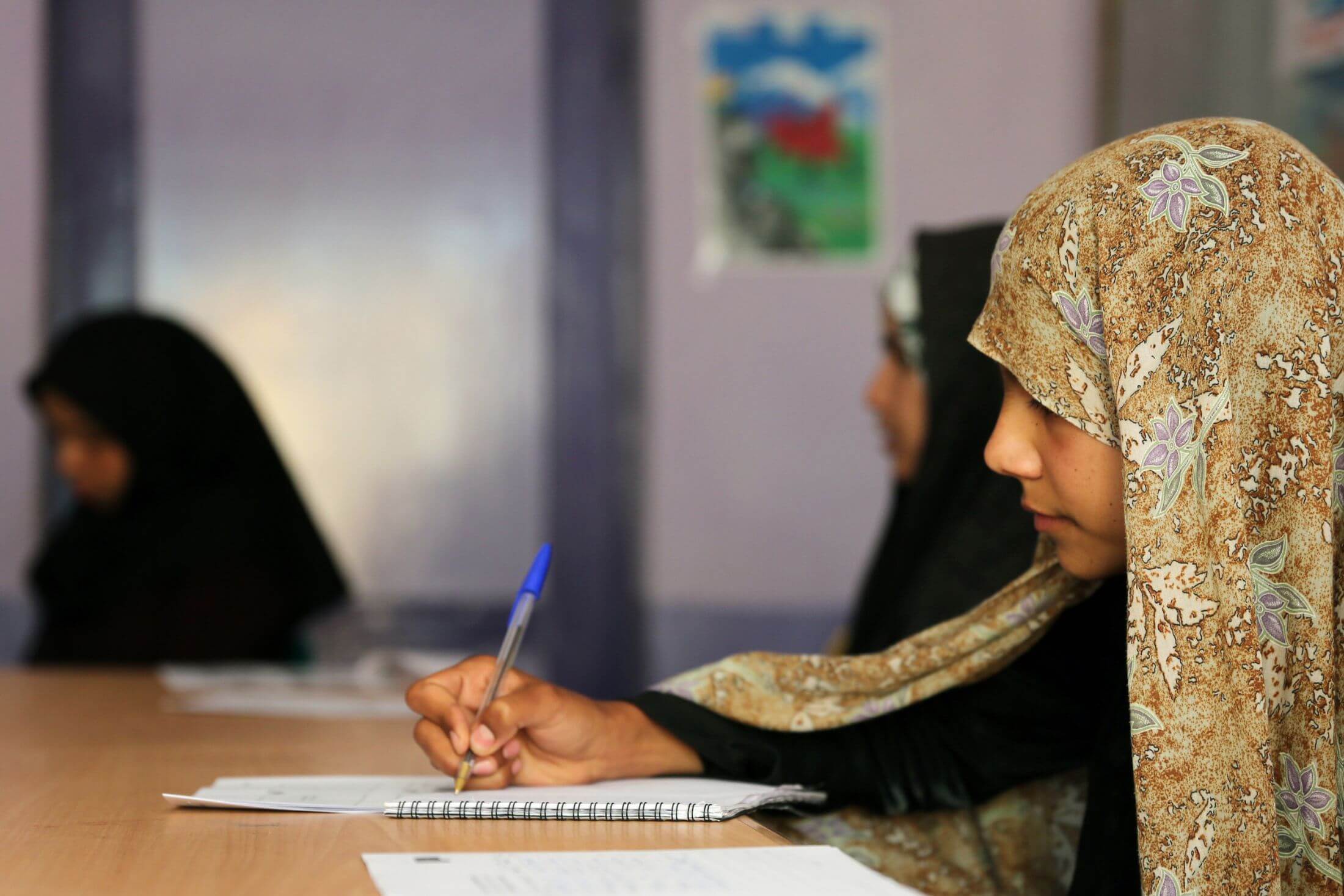 Young woman in a headscarf sits at a table in a classroom with two other young women in headscarves and writes in a notebook