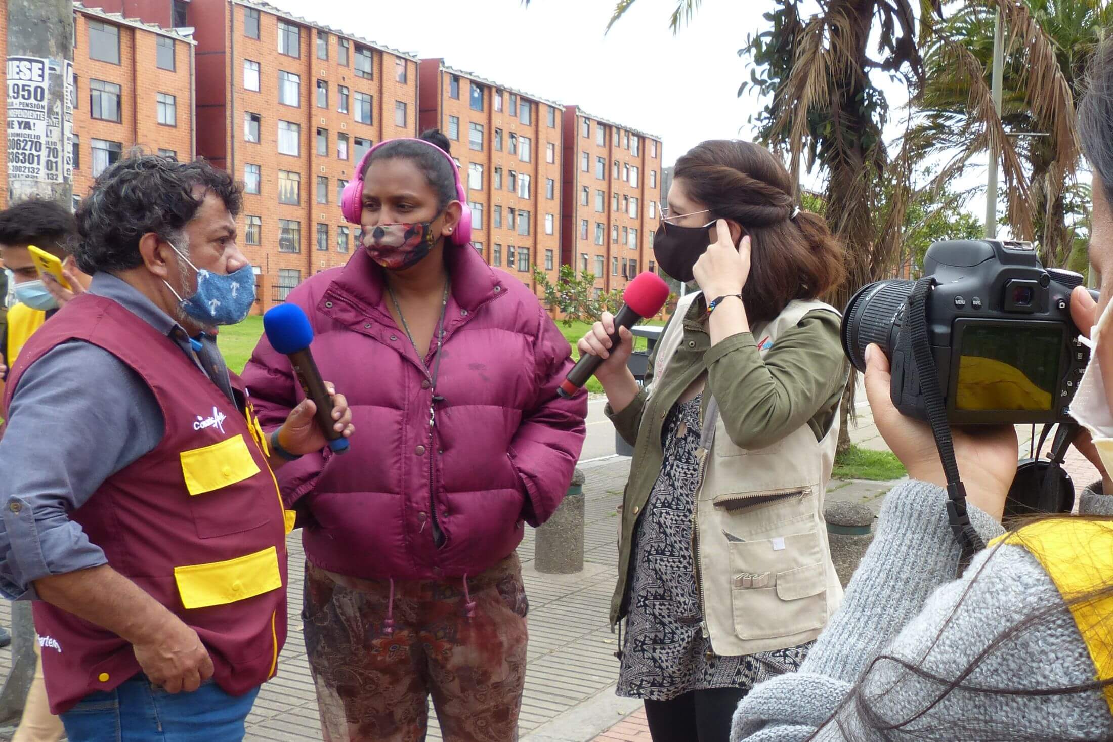 A male and a female citizen journalist interview a woman outside in a public square while a woman videos with a camera. All are wearing face masks.