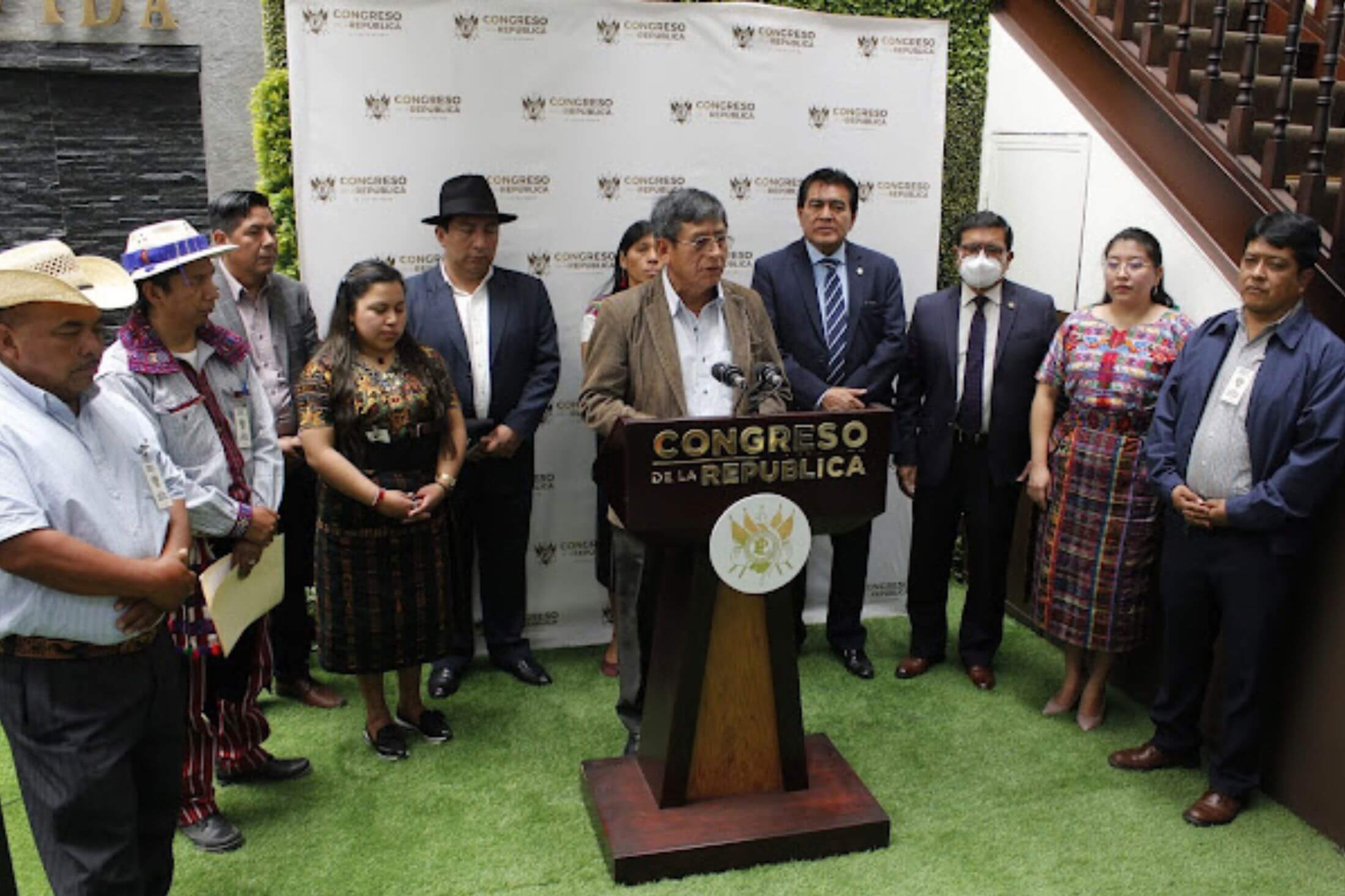 An Indigenous man speaks at a podium at a press conference at the Guatemalan Congress. Ten Indigenous people stand in a semi-circle behind him.
