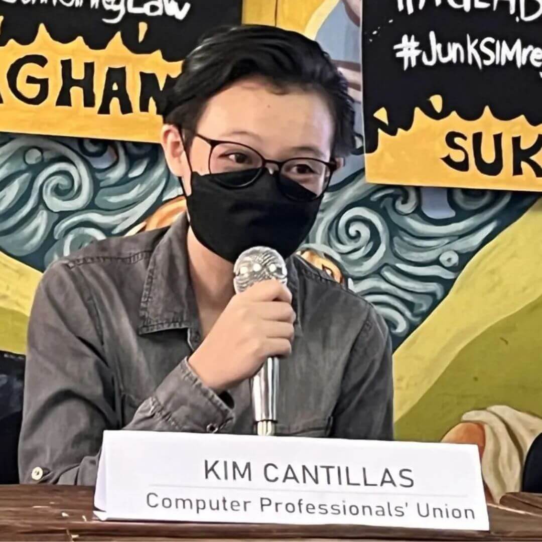 A younger-looking person wearing glasses and a black facial mask sits at a table and speaks into a microphone. A name card in front of them says Kim Cantillas, Computer Professionals' Union. Behind is a black, yellow and grey-colored banner with writing in Filipino.