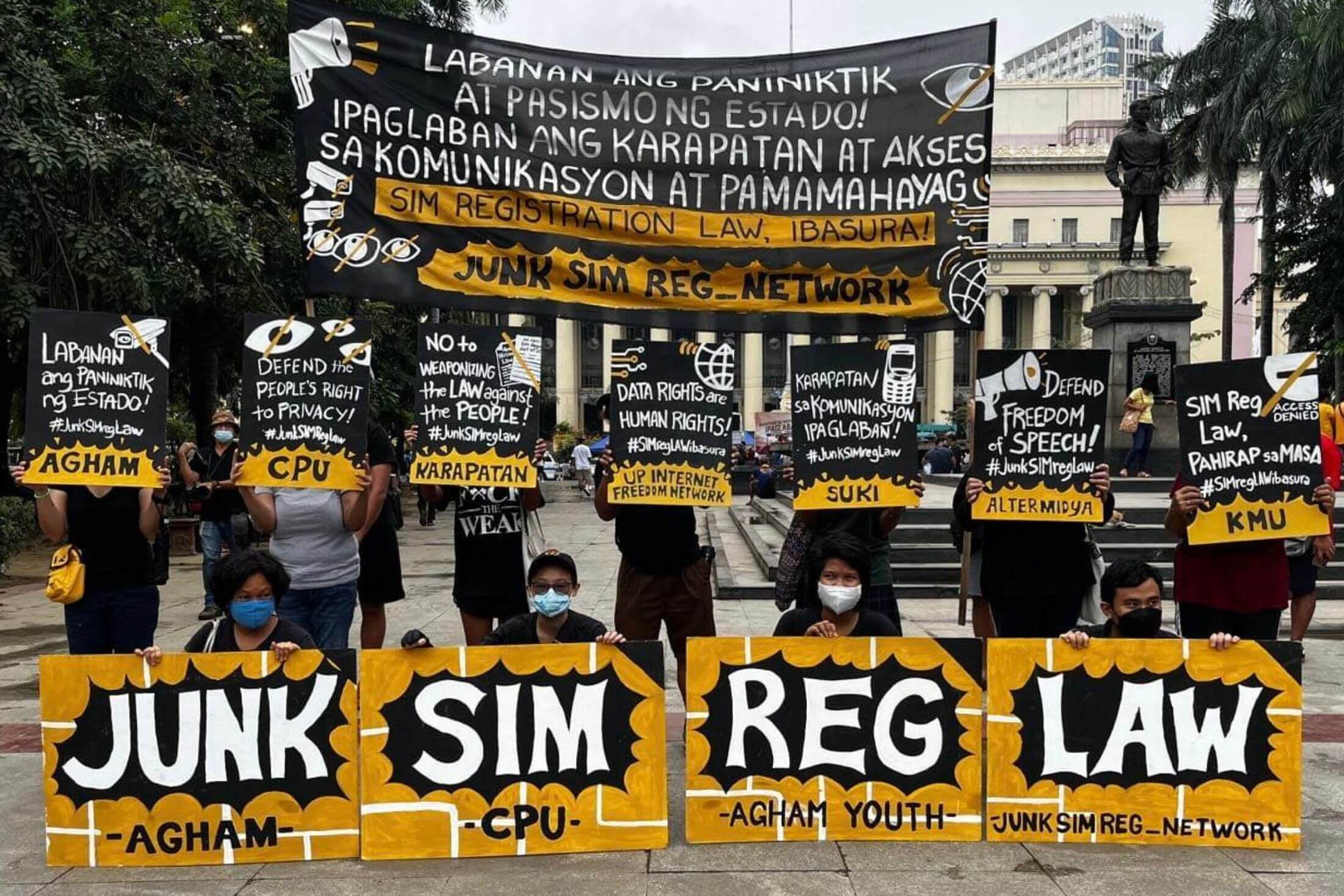 Eleven people hold black and yellow signs with white slogans written on them in Filipino and English at a demonstration in an outside plaza. Four of the people are kneeling and seven are standing behind them.
