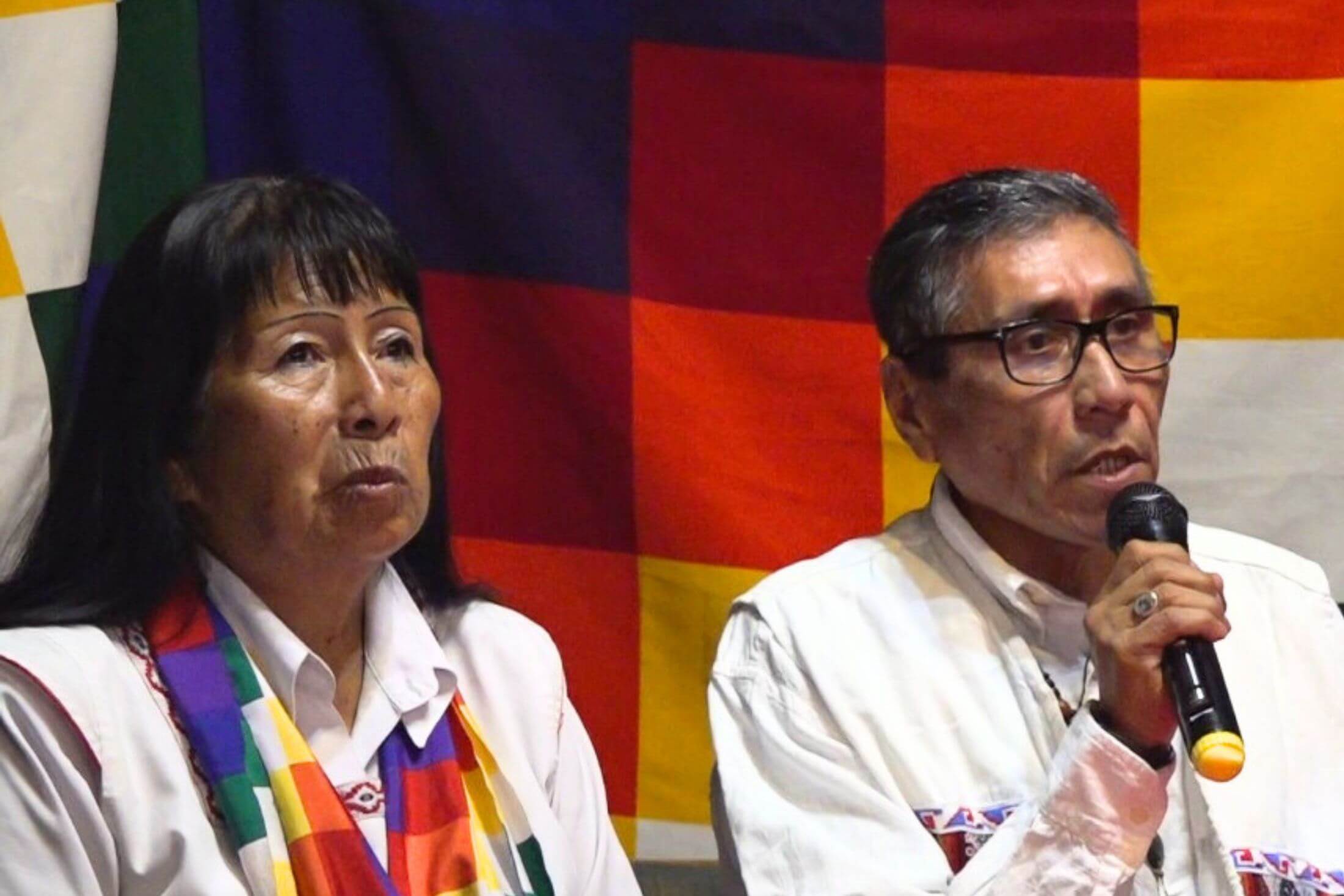 Indigenous woman and man from Argentina, wearing white tops. The man is speaking into a microphone. Behind them is the Wiphala Indigenous flag made up of squares coloured dark brown, dark red, orange, yellow and white. The woman wears a scarf with the same emblem.